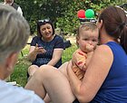 Eine Frau sitzt im Park auf der Wiese und unterhält sich mit Eltern, im Vordergrund eine Mutter mit Kleinkind im Arm