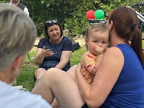 Eine Frau sitzt im Park auf der Wiese und unterhält sich mit Eltern, im Vordergrund eine Mutter mit Kleinkind im Arm