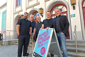 Fünf Männer stehen vor dem Theatergebäude an einem Aufsteller mit der Beschriftung "Der neue Spielzeitkalender 23/24"